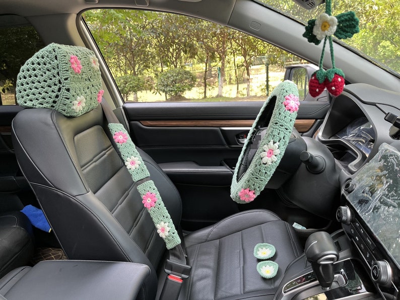 May include: Green crocheted car seat covers with pink flowers. The set includes a headrest cover, seatbelt cover, and steering wheel cover.