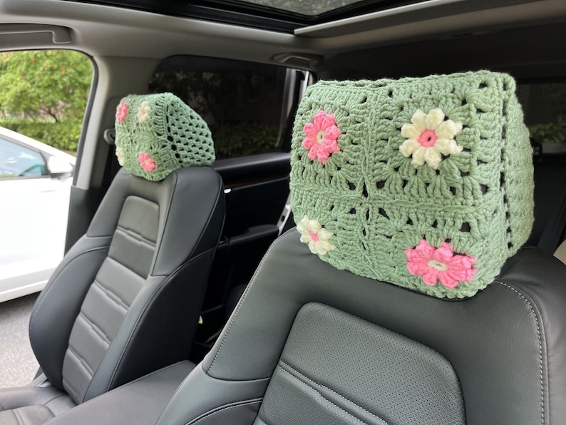 May include: Two crocheted headrest covers with a green background and pink and white flowers. The covers are placed on the headrests of two car seats.
