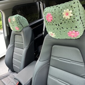 May include: Two crocheted headrest covers with a floral design in shades of green, pink, and white. The covers are placed on the headrests of two car seats.