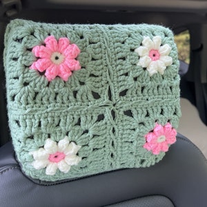 May include: A green crocheted headrest with pink and white flowers. The headrest is made of a soft, textured yarn and is perfect for adding a touch of comfort and style to any car.