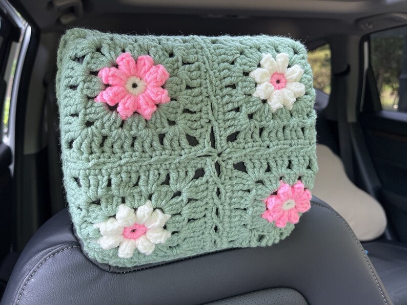 May include: A green crocheted headrest with pink and white flowers. The headrest is made of a soft, textured yarn and is perfect for adding a touch of comfort and style to any car.