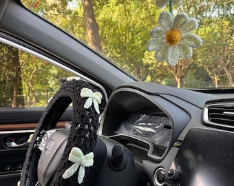 May include: A black crochet steering wheel cover adorned with light green bows. Two crochet daisy car air fresheners hang from the rearview mirror. The interior of a car is visible, with a view of trees through the windshield.