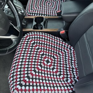 May include: Crocheted car seat covers in a black, white, gray, and red pattern. The covers are placed on the front seats of a car, with the steering wheel and console visible. The crochet design features a repeating pattern of small, colorful squares.