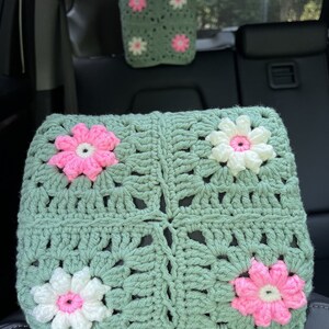 May include: A green crocheted car seat headrest with pink and white flowers. The headrest is made of a soft, textured yarn and has a square shape.