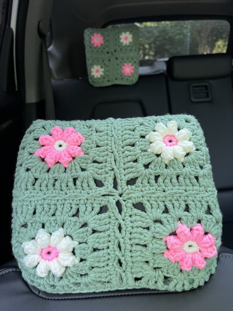 May include: A green crocheted car seat headrest with pink and white flowers. The headrest is made of a soft, textured yarn and has a square shape.
