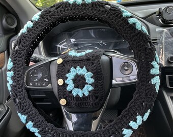 Peut inclure: Housse de volant en crochet noir avec des accents de fleurs bleues. La housse est sur un volant en cuir noir dans une voiture.