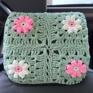May include: A green crocheted headrest with pink and white flowers. The headrest is made of a soft, textured yarn and has a square shape.