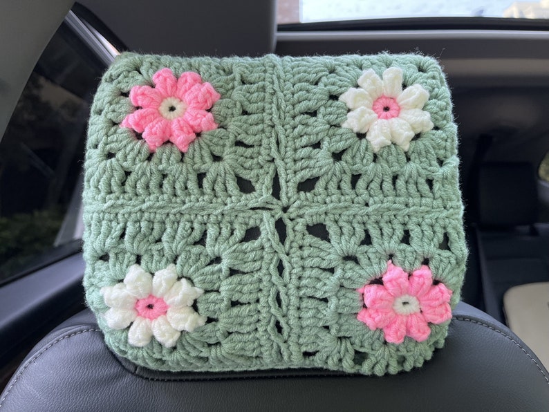 May include: A green crocheted headrest with pink and white flowers. The headrest is made of a soft, textured yarn and has a square shape.