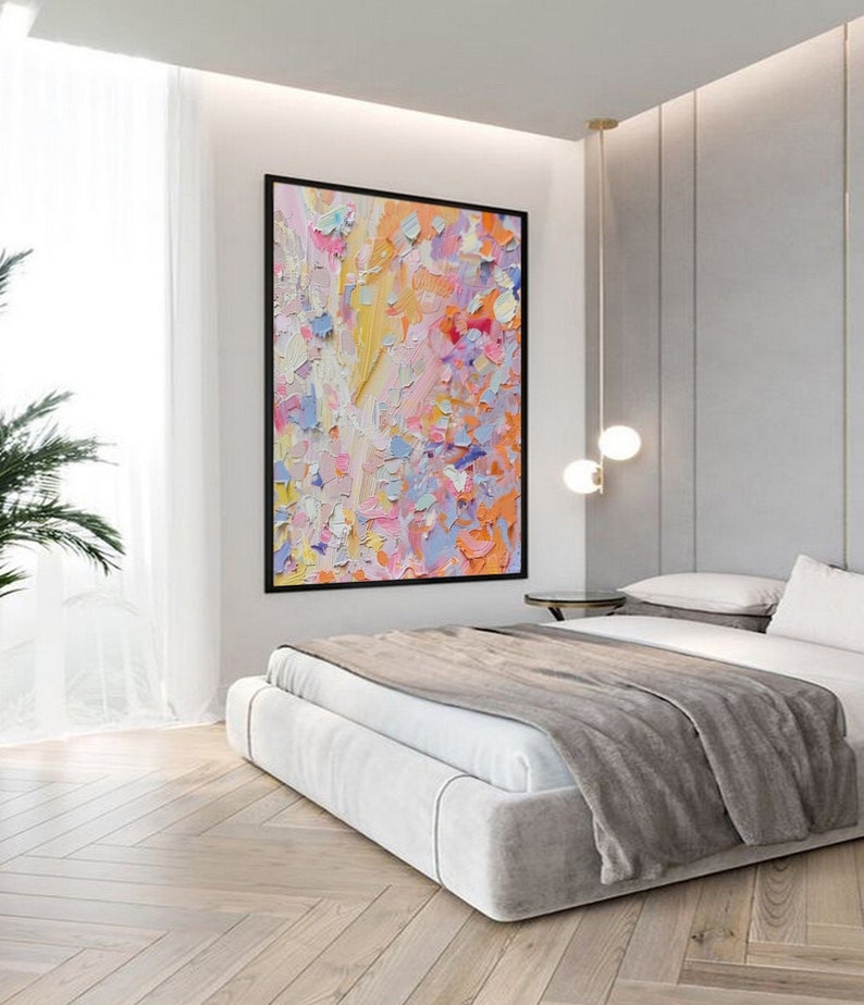 May include: A large abstract painting with a black frame hangs above a bed. The artwork features a textured, colorful palette of orange, pink, yellow, and blue. The bedroom has a modern design with a light-colored bed and wooden floors.