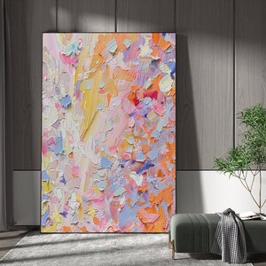May include: Abstract art painting with a textured surface. The artwork features a vibrant mix of colors, including pink, orange, yellow, and blue. The painting is displayed in a black frame, leaning against a wooden wall.