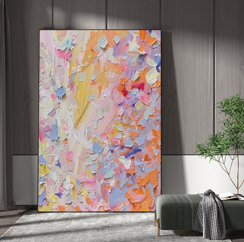 May include: Abstract art painting with a textured surface. The artwork features a vibrant mix of colors, including pink, orange, yellow, and blue. The painting is displayed in a black frame, leaning against a wooden wall.