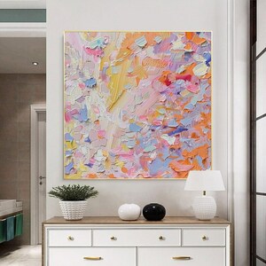 May include: Abstract oil painting with a gold frame. The artwork features a textured surface with a palette of colors including yellow, pink, orange, blue, and white. The painting is displayed above a white dresser.
