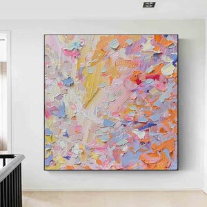 May include: Abstract oil painting with a textured surface. The artwork features a vibrant palette of colors, including yellow, pink, orange, and blue. The painting is displayed in a black frame, adding a modern touch to the colorful composition.