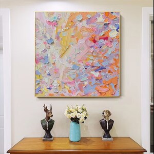 May include: Abstract oil painting in a gold frame, featuring a textured surface with a palette of pink, orange, yellow, and blue hues. The artwork hangs above a wooden cabinet with decorative busts and a vase of white flowers.