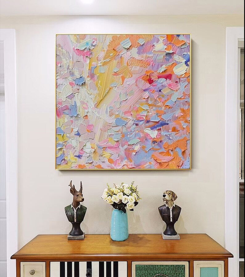 May include: Abstract oil painting in a gold frame, featuring a textured surface with a palette of pink, orange, yellow, and blue hues. The artwork hangs above a wooden cabinet with decorative busts and a vase of white flowers.