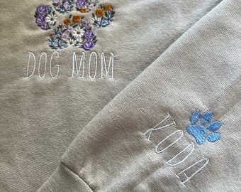 May include: A tan sweatshirt with embroidered floral paw print design and the words "DOG MOM". The sleeve cuff features the name "KODA" and a blue paw print.