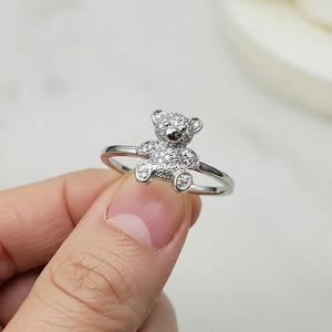 May include: A silver ring featuring a small teddy bear design. The bear is covered in small, sparkling crystals. The ring is held up by a person's fingers, showcasing the ring's delicate details and overall design.