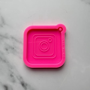 May include: Bright pink silicone mould in the shape of the Instagram logo. The mould has a square outline with rounded corners and a small loop at the top. The Instagram logo is embossed within the square. The text "ANGEL WINGS" is printed on the side.