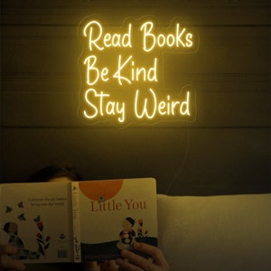 May include: A yellow neon sign with the words "Read Books Be Kind Stay Weird" on a dark brown wall. A person is reading a book titled "Little You" in the foreground.