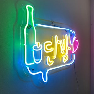 May include: Neon sign with a blue, green, and yellow outline. The sign says "다방" in Korean, which means "coffee shop". There is a stylized image of a bottle and a cup on the sign.
