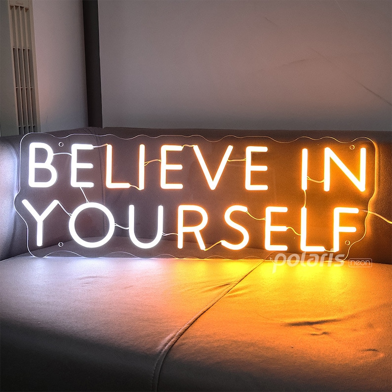 May include: White neon sign with the words "Believe in Yourself" in a wavy font. The sign is lit up with a warm yellow glow.