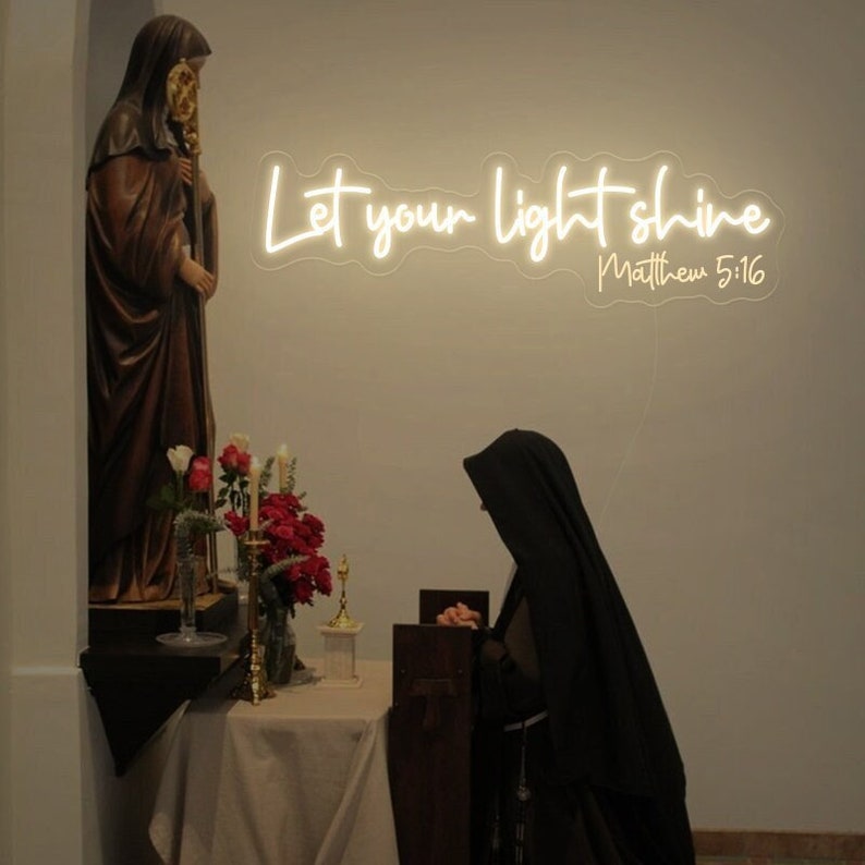May include: A neon sign with the words "Let your light shine" and the reference "Matthew 5:16" in a church setting. A statue and a person in a black habit are in the foreground. Red and white flowers are in vases.
