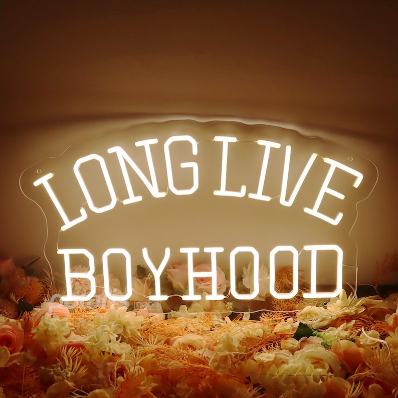May include: White neon sign with the text "LONG LIVE BOYHOOD" on a clear acrylic background. The sign is sitting on a bed of pink and white flowers.