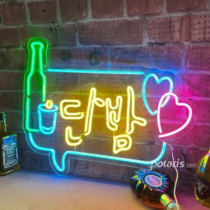 May include: Neon sign with a speech bubble shape, featuring a green bottle, a yellow candle, and the Korean text "Let's Drink" in yellow. Two pink hearts are on the right side of the sign.