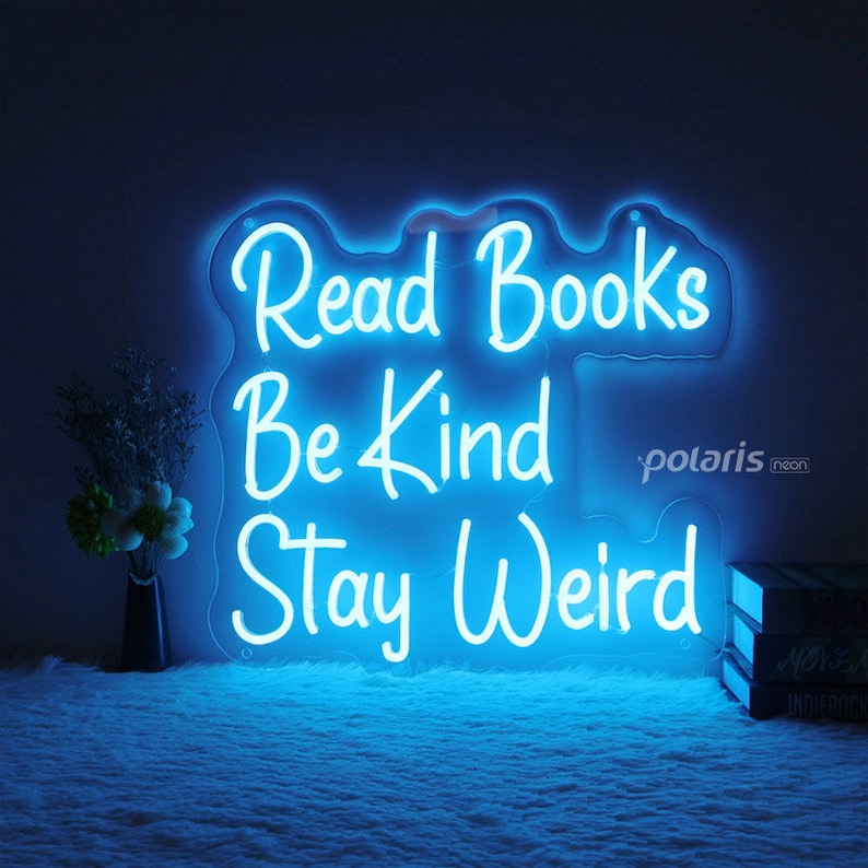 May include: Blue neon sign with the words "Read Books Be Kind Stay Weird" on a white background. The sign is lit up and is hanging on a wall.