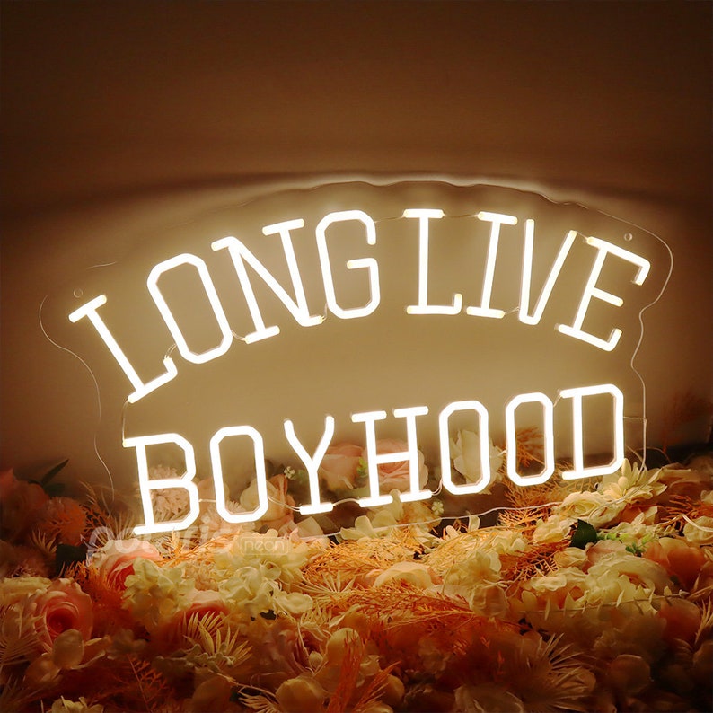 May include: White neon sign with the words "Long Live Boyhood" on a clear acrylic background. The sign is sitting on a bed of artificial flowers.