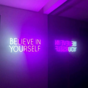 May include: Two neon signs with the phrase "Believe in Yourself" in white and pink on a purple wall.