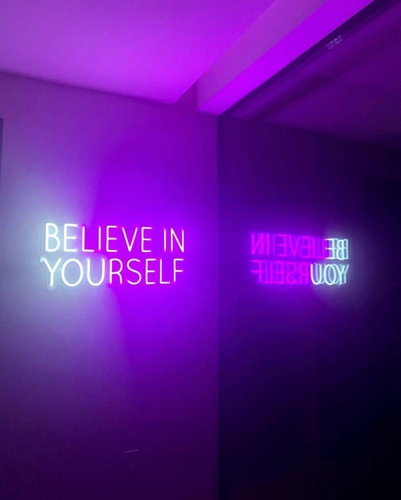 May include: Two neon signs with the phrase "Believe in Yourself" in white and pink on a purple wall.