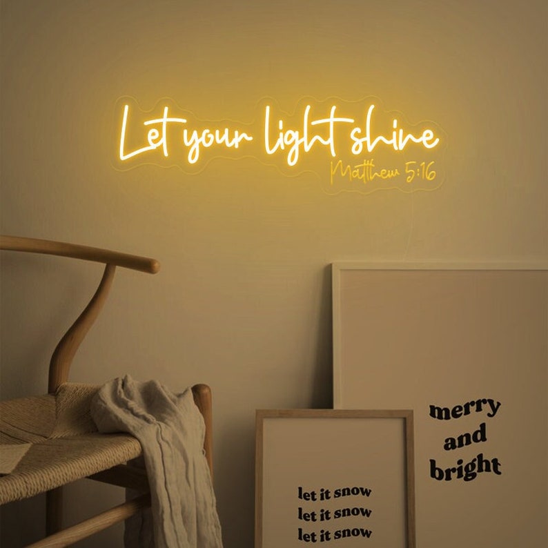 May include: A neon sign with the words "Let your light shine" in a cursive font, with "Matthew 5:16" below. The sign is illuminated in a warm yellow color. The image also includes a wooden chair, a framed print, and a print with the words "merry and bright".