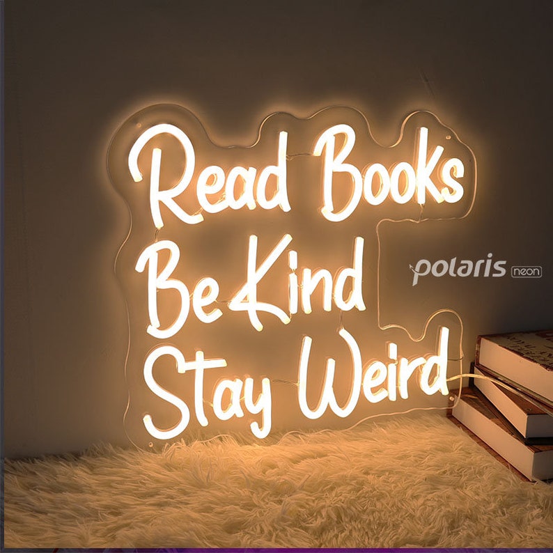 May include: White neon sign with the words "Read Books Be Kind Stay Weird" on a white background. The sign is lit up and is hanging on a wall.