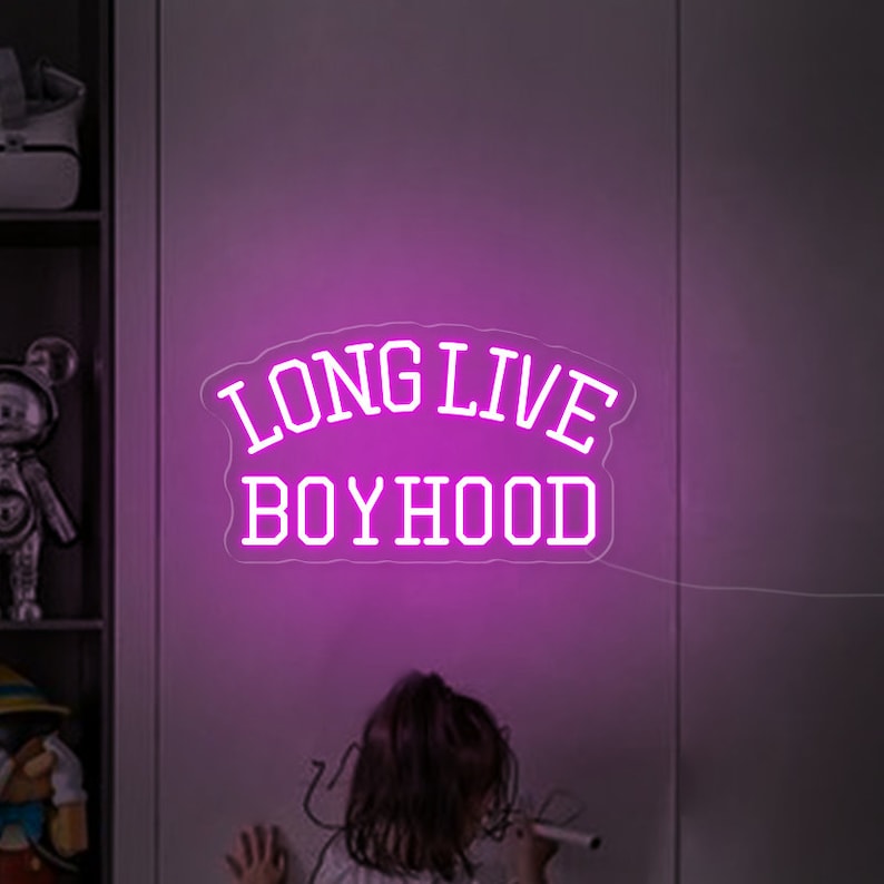 May include: Pink neon sign with the text "LONG LIVE BOYHOOD" in white lettering. The sign is mounted on a gray wall.