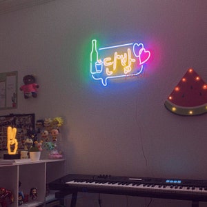 May include: A neon sign with the Korean phrase "Let's drink" in a yellow speech bubble with a bottle and glass of alcohol. The sign is lit up in rainbow colors and is mounted on a white wall. There is a watermelon-shaped light on the wall to the right of the sign.
