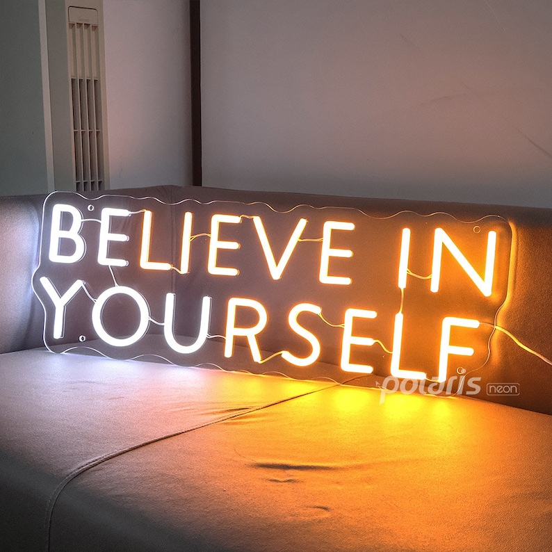May include: White neon sign with the words "Believe in Yourself" in a wavy font. The sign is lit up with a warm yellow glow.