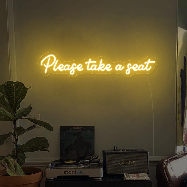 May include: A yellow neon sign with the text "Please take a seat" on a wall. The sign is lit up and is hanging above a record player and a small amplifier.