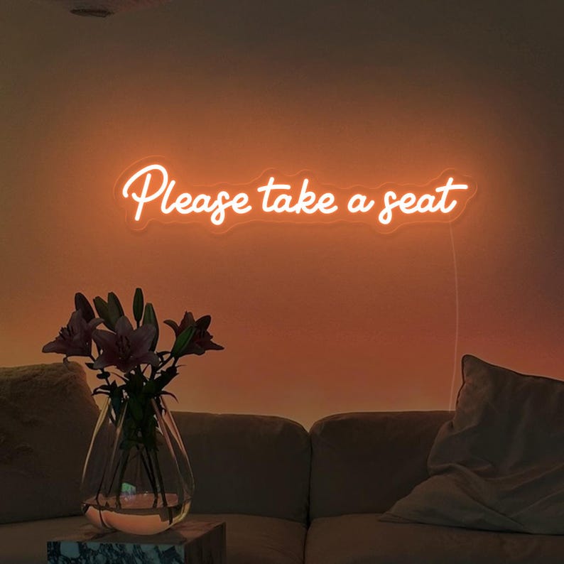 May include: A neon sign with the text "Please take a seat" in white letters on an orange background. The sign is mounted on a wall above a couch with a throw pillow.