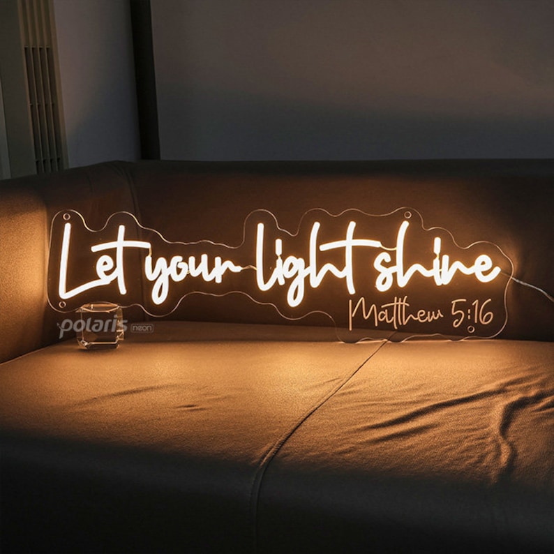 May include: A neon sign with the words "Let your light shine" and the reference "Matthew 5:16" in a flowing script. The sign is illuminated in a warm white color and mounted on a clear backing.