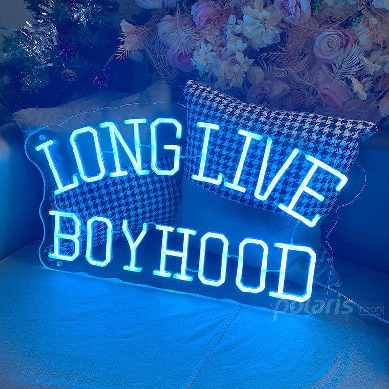 May include: A blue neon sign with the words "Long Live Boyhood" in a white outline. The sign is on a blue couch with a black and white patterned pillow.