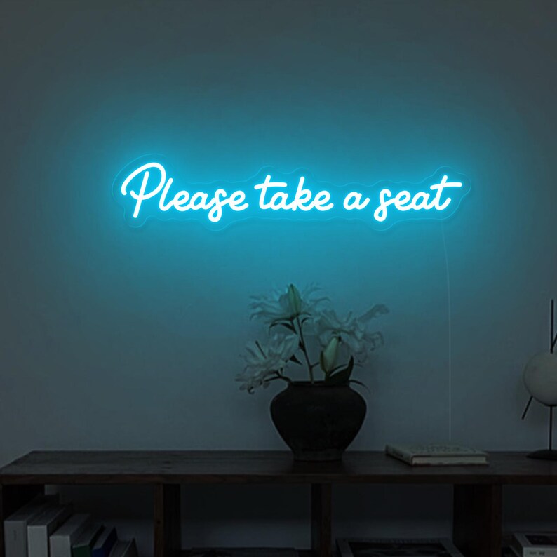 May include: A blue neon sign with the text "Please take a seat" on a white wall. The sign is hanging above a wooden shelf with a vase of white flowers and a book.