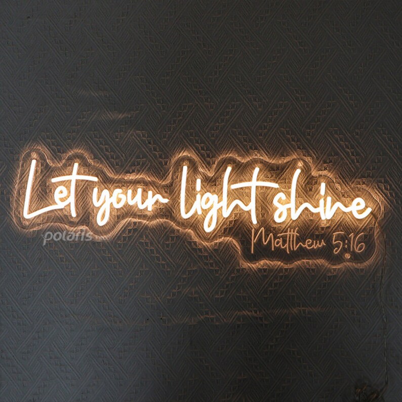 May include: A glowing neon sign with the words "Let your light shine" in a cursive font. Below the text is "Matthew 5:16". The sign is a warm, inviting color and is mounted on a textured gray wall.