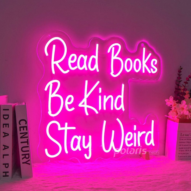 May include: Pink neon sign with the text "Read Books Be Kind Stay Weird" on a clear acrylic background.