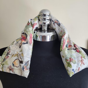 Peut inclure: Un tour de cou en tissu crème orné d'un motif imprimé de papillons colorés, de petits oiseaux, de baies rouges et de champignons. Le tissu présente un motif de fond en forme de grille et est drapé sur un présentoir métallique.