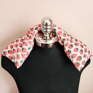 May include: A neck wrap with a white background and a pattern of red strawberries with green leaves. The wrap is draped over a black mannequin. The wrap is designed to be worn around the neck and shoulders.