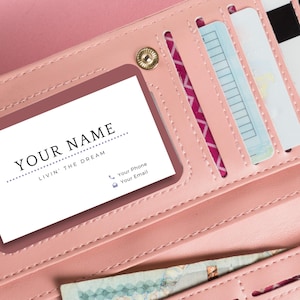 Puede incluir: Una cartera de cuero rosa con una ranura para tarjeta que dice "YOUR NAME LIVIN' THE DREAM" y un icono de tel&eacute;fono y correo electr&oacute;nico. La cartera tiene varias ranuras para tarjetas y un bolsillo para dinero.