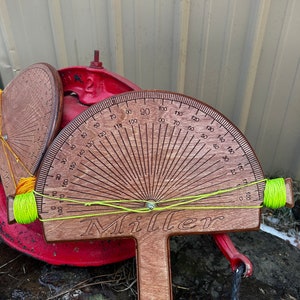 May include: Wooden protractor with a semi-circular scale, marked from 0 to 180 degrees, and the name "Miller" engraved. The tool has a T-shaped handle and is strung with neon green thread. A smaller, similar wooden disc is also visible.