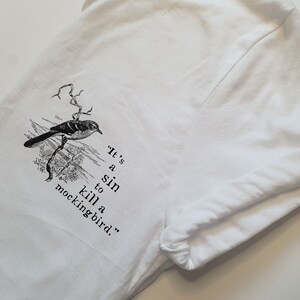 May include: White t-shirt featuring a black and white illustration of a bird on a branch. The text "It's a sin to kill a mockingbird." is printed below the bird.