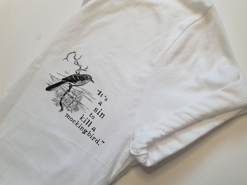 May include: White t-shirt featuring a black and white illustration of a bird on a branch. The text "It's a sin to kill a mockingbird." is printed below the bird.